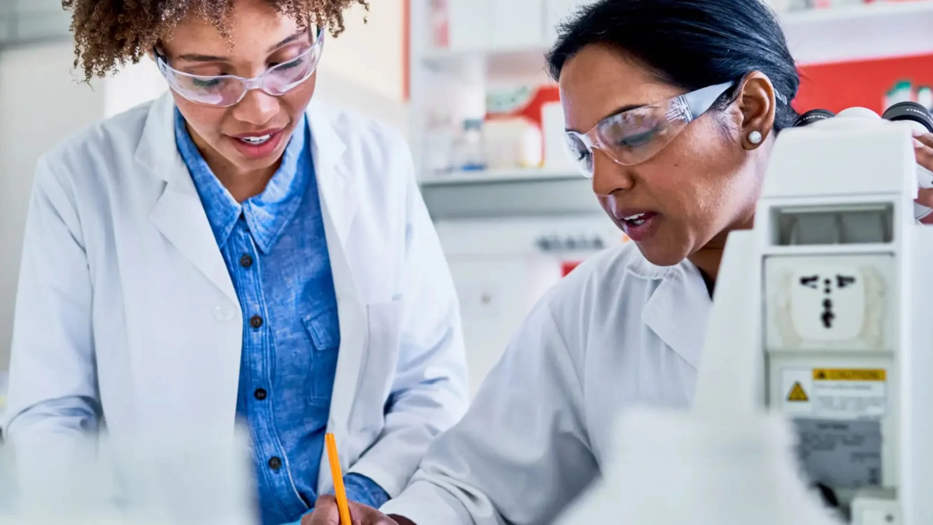 Two scientists wearing safety glasses and lab coats reviewing research notes in a modern laboratory beside scientific equipment.