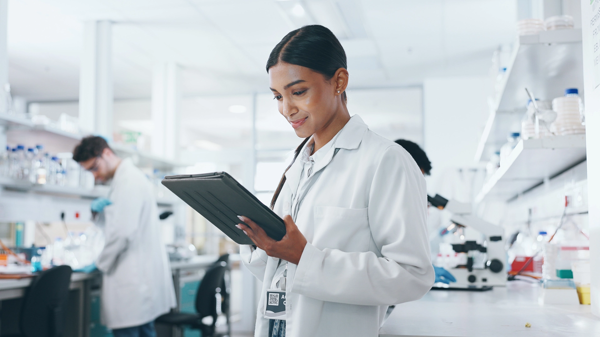 Scientist in a modern laboratory using a digital tablet while colleagues conduct research in the background.